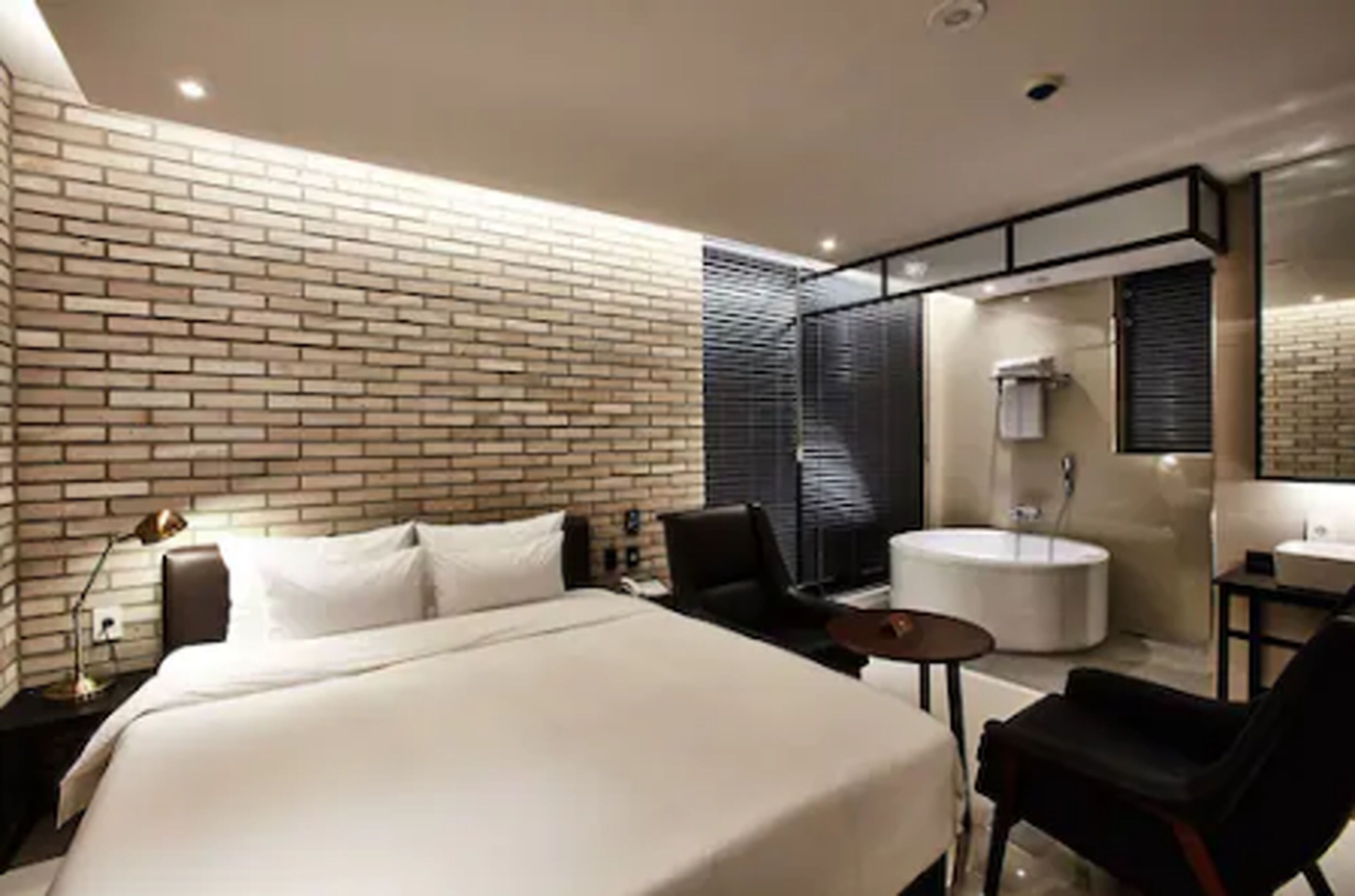 Een moderne hotelkamer met een bakstenen muur, een groot bed, een ronde spiegel, een klein tafeltje, een stoel en een badkamer met bad en wastafel.