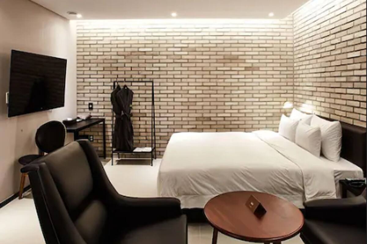 Une chambre d’hôtel moderne avec un mur au motif de briques, un téléviseur à écran plat, un lit avec du linge de maison blanc, une petite table ronde et un fauteuil en cuir noir.