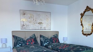 Ein Schlafzimmer mit einem großen Bett, einer Kristallleuchtre, einem gerahmten Blumenbild und einem Spiegel mit aufwendiger Rahmenornierung.
