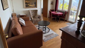 Ein Wohnzimmer mit Sofa, Sessel, Esstisch, einer Chandelieber und einem Gemälde an der Wand.