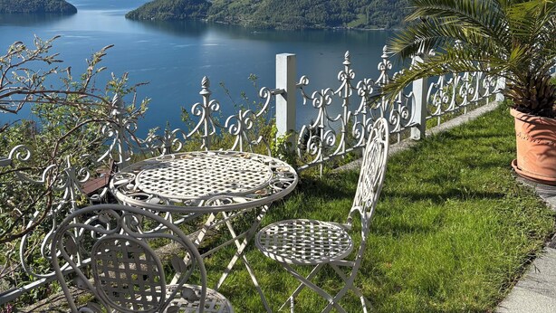 Eine Terrasse mit Metallstühlen und einem Tisch, mit Blick auf einen See und Berge.