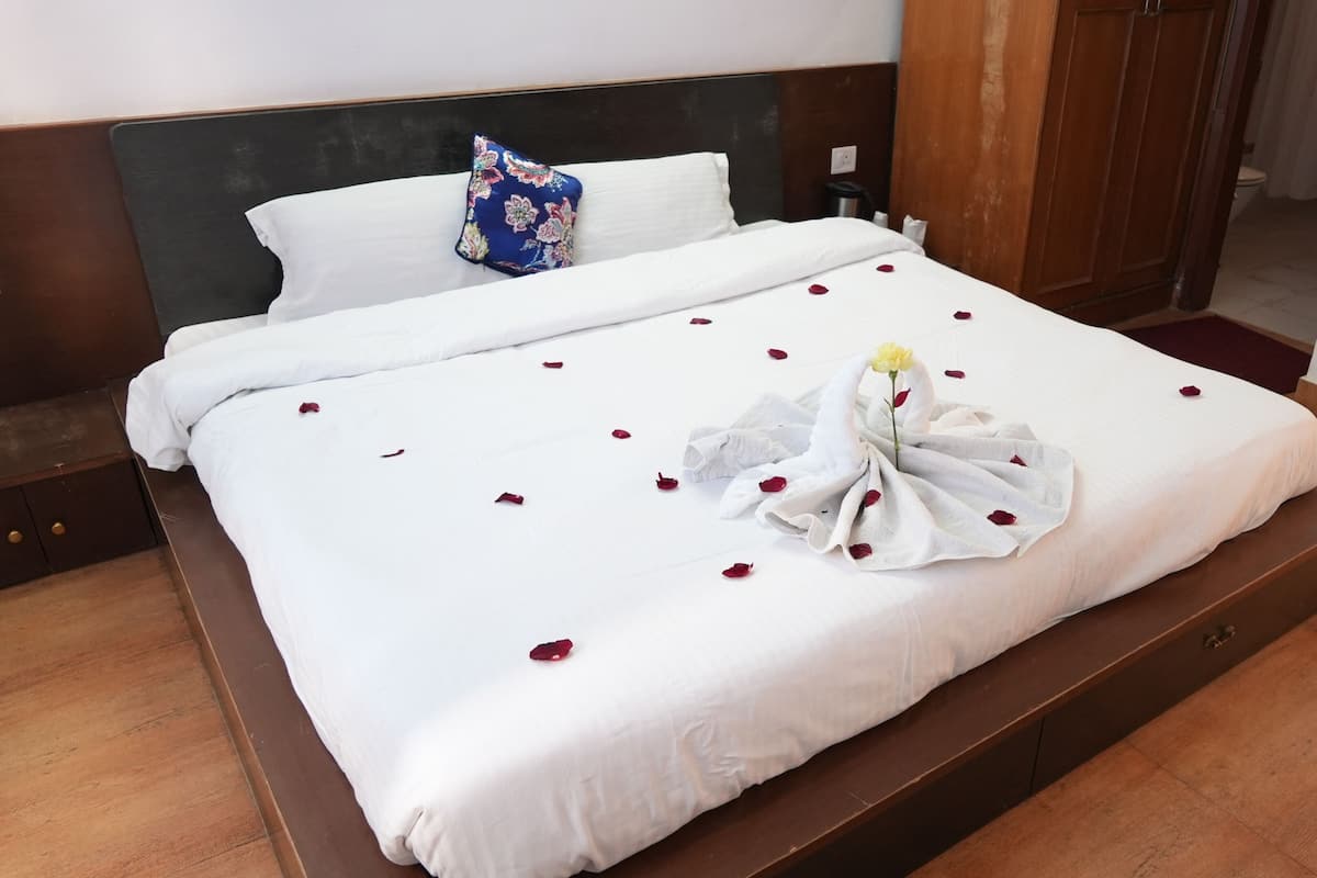 Una cama bien arreglada con sábanas blancas, un cojín floral azul y pétalos de rosa esparcidos sobre la cama.