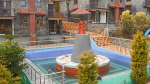 A swimming pool with a central fountain, surrounded by stone buildings with red roofs and balconies. - Nature Bloom Dharamshala Hotel and Resort (Palampur)