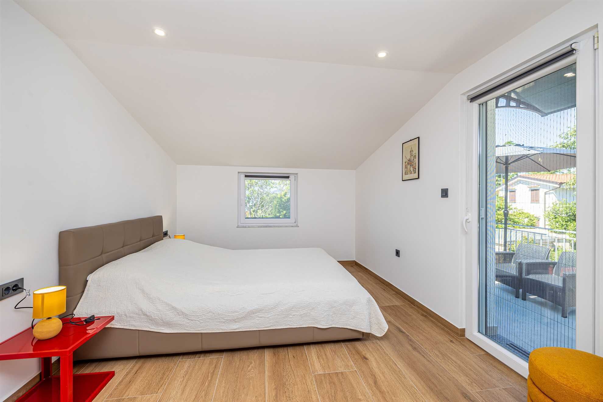 Een slaapkamer met een bed, een klein raam, een rode tafel en een balkon met tuinmeubilair.