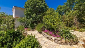 A stone pathway leading to a building with a tiled roof, surrounded by lush greenery and flowering plants. - Holiday house MICHAEL with shared pool and private jacuzzi (Pinezići)