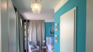 Een kamer met een houten vloer, een grijze fauteuil met een blauw kussen, een nachtkastje met een lamp, een bed met gestreept beddengoed en een deur met een handgreep.
