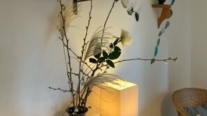 Et værelse med et træbord, en vase med grene, en lampe og en fletstol.
