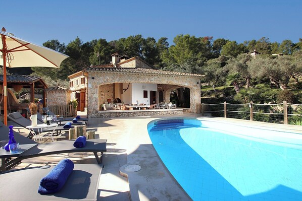 A poolside area with lounge chairs, a covered seating area, and a stone building with a tiled roof. - Finca Lourdes, quiet location, pool, air conditioning, 5 bedrooms, 5 bathrooms (Pollença)