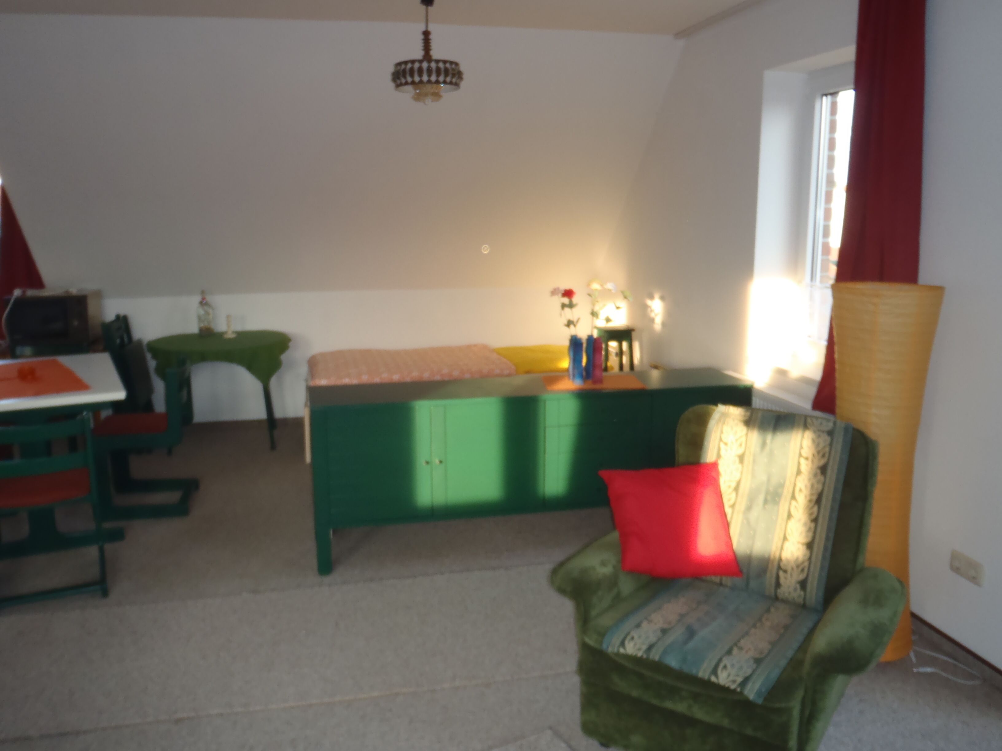 A room with a green sofa, a small table with a pink cushion, a green cabinet, and a window with red curtains.