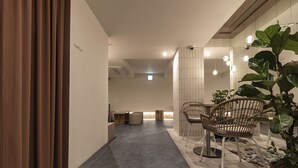 A hallway with a modern design, featuring a seating area with chairs and a table, a potted plant, and a sign indicating the direction to the toilet. - Tong Tong Petit Hotel (Seoul)