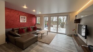 Un salon moderne avec un mur d’accent rouge, un canapé en cuir, une table basse en bois et une grande porte-fenêtre coulissante donnant sur une terrasse.
