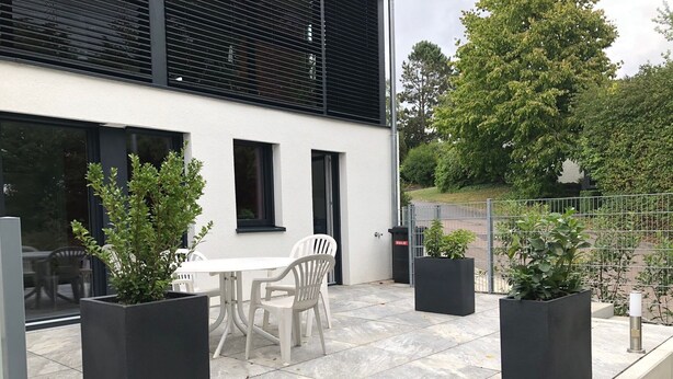 Un espace patio extérieur avec une table et des chaises blanches, entouré de jardinières noires contenant des plantes vertes.