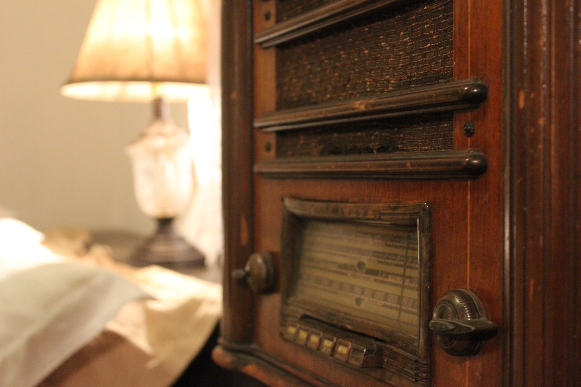 A wooden radio with a dial and buttons, a lamp, and a bed with a white sheet.