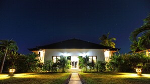 A two-story building with a central entrance, surrounded by a well-maintained garden with palm trees and a paved pathway leading to the entrance. - Authentic Beachvilla with cook, private pool, staff and lush tropical garden (Seririt)