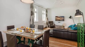 Un comedor con mesa de vidrio, sillas, alimentos para el desayuno y vista a una sala de estar con sofá y un cuadro.