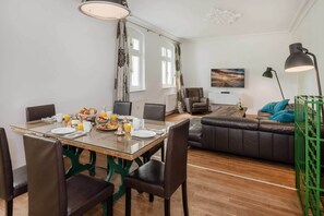 Un coin repas avec une table en verre, des chaises, des aliments pour le petit-déjeuner et une vue sur un salon comprenant un canapé et un tableau.