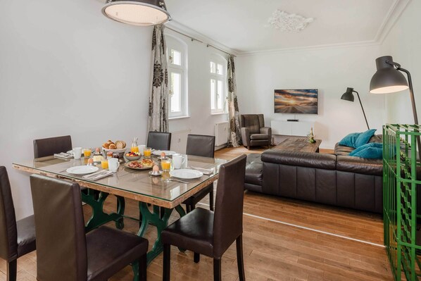 Ein Essbereich mit einem Glastisch, Stühlen, Frühstücksangeboten und Blick auf ein Wohnzimmer mit einer Couch und einem Gemälde.