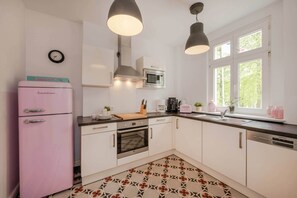 Une cuisine avec des meubles blancs, un réfrigérateur rose, un four à micro-ondes, une cuisinière et un évier. On y trouve deux lampes suspendues, une horloge et une fenêtre donnant sur un paysage verdoyant.