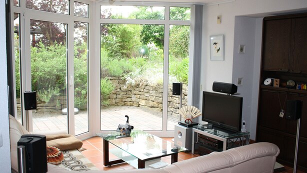 A living room with a glass coffee table, a beige sofa, a TV, and a stone wall visible through the sliding glass doors.