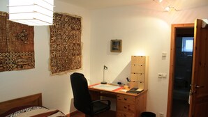 A bedroom with a bed, a desk, a chair, a wooden cabinet, a trash bin, and a door leading to a bathroom.