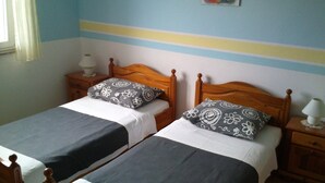 A room with two single beds, each with a wooden headboard and a bedside table with a lamp. The walls have a striped pattern.