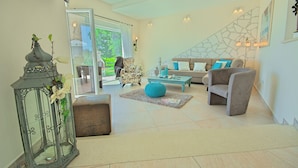 Un salon spacieux avec un canapé, des fauteuils et une table basse. On y trouve une lanterne décorative et, grâce à une porte vitrée, on peut admirer la vue sur l’extérieur.