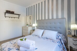 Ein ordentlich bezogenes Bett mit Blumendekor, ein Nachttisch mit Lampe und ein an der Wand befestigtes Regal mit Körben.