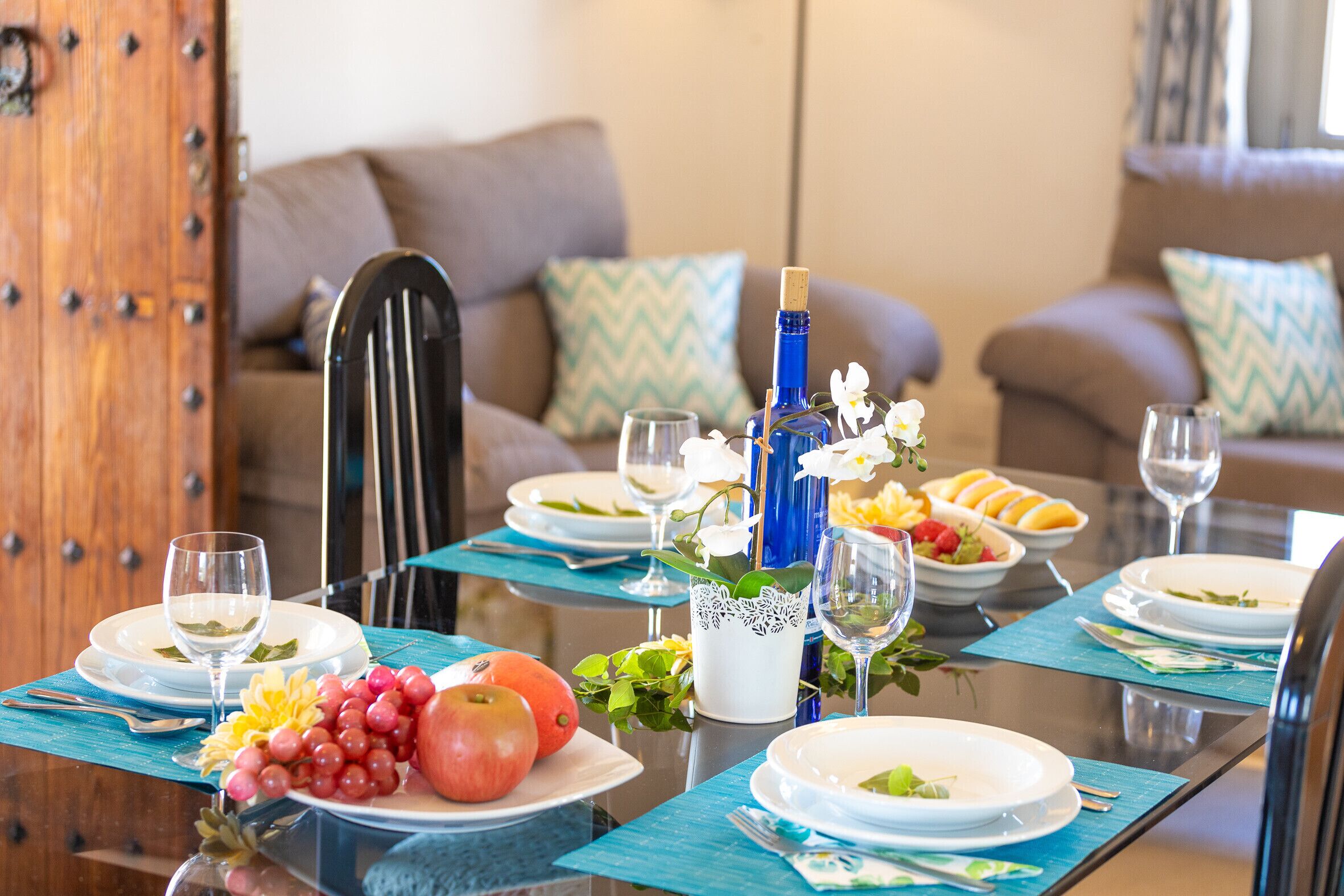 Una mesa de comedor dispuesta con platos, vasos y una botella, rodeada de sillas y un sofá en una habitación acogedora.