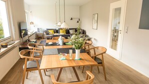 A modern dining area with a wooden table and chairs, a vase with flowers, and a view of the living room in the background. - Feha - Holiday House "Minnet", 15169 (Leer (Ostfriesland))