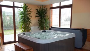 A hot tub in a room with large windows, a wooden bench, and potted plants.