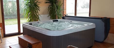 A hot tub in a room with large windows, a wooden bench, and potted plants.