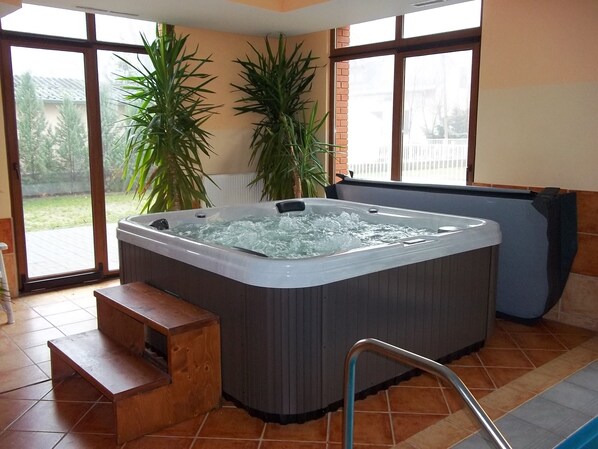 A hot tub in a room with large windows, a wooden bench, and potted plants.
