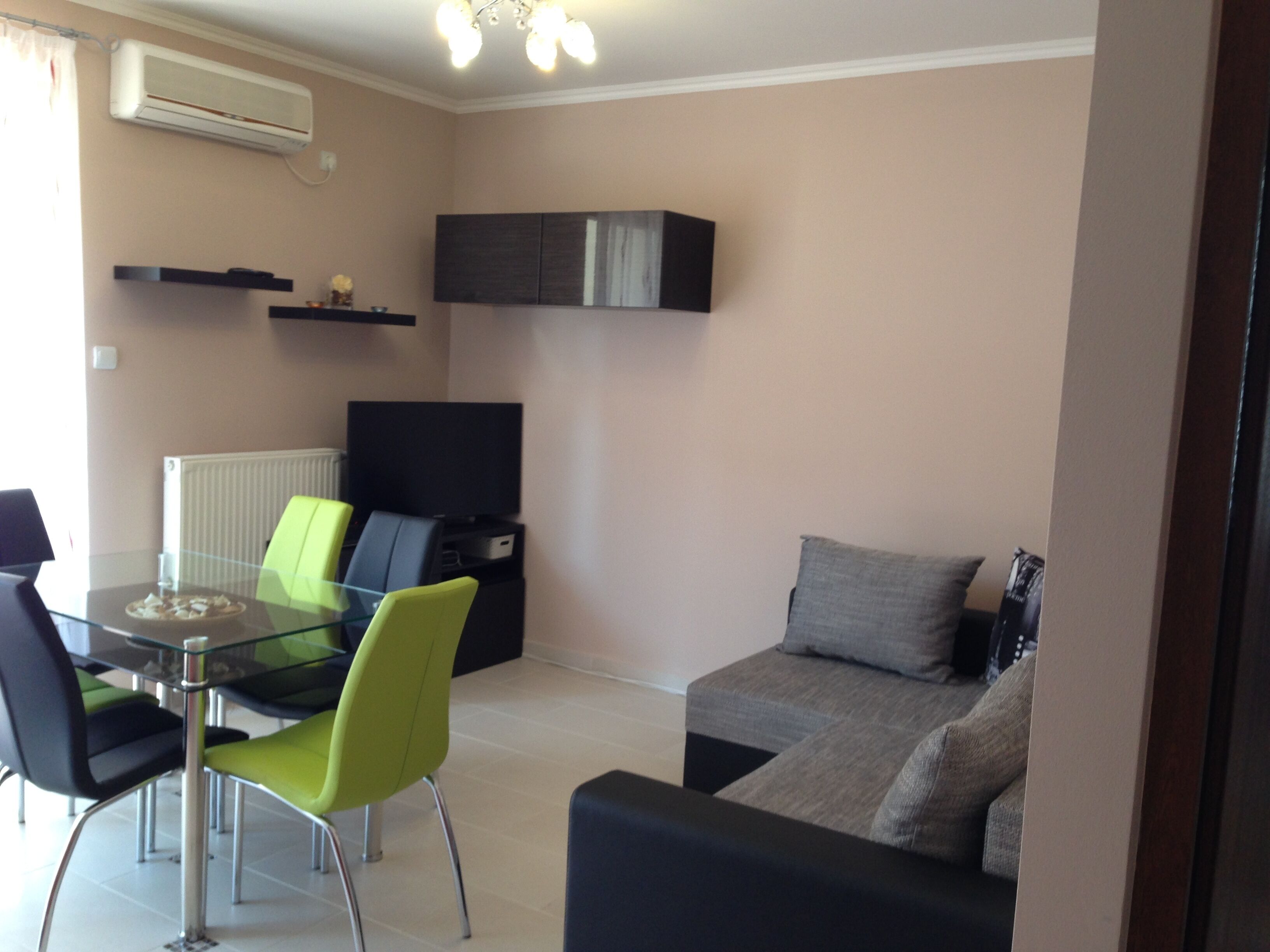 A modern living room with a glass dining table, green and black chairs, a grey sofa, and a flat-screen TV.