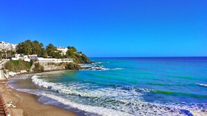 A beach with waves, a clear blue sky, and buildings in the background.