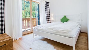 Un dormitorio con cama blanca, suelo de madera, alfombra mullida blanca y vistas a una terraza de madera y árboles a través de una puerta de vidrio.