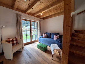 Una habitación con techo de madera, un sofá con cojines cuadriculados, una silla con un cojín escocés, un taburete pequeño y una caja verde en el suelo.