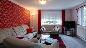 Una sala de estar con un sofá, un sillón adicional, una mesa de centro de vidrio, un televisor y un radiador.