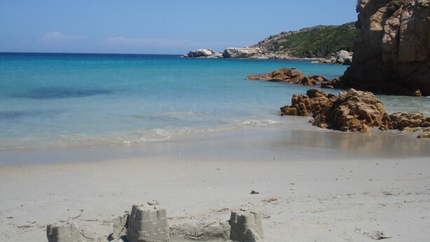 A sandy beach with clear blue water, rocky formations, and a mountainous backdrop.
