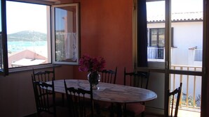 A dining area with a table and chairs, a vase with flowers, and large windows with views of the sea and buildings.