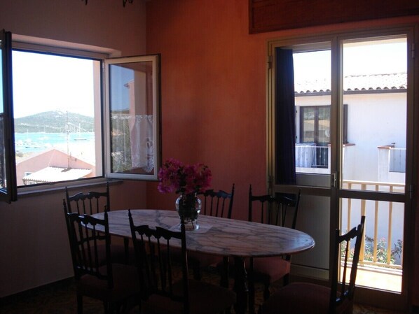 A dining area with a table and chairs, a vase with flowers, and large windows with views of the sea and buildings.