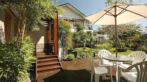 A patio with wooden steps leading to a white chair and table under a large umbrella, surrounded by lush greenery and a modern house. - Kanazawa House (Ji'an)