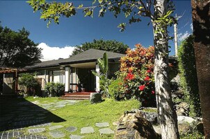 Una casa con porche cubierto, un camino de piedra que conduce a la entrada y un jardín con diversas plantas y un árbol grande.