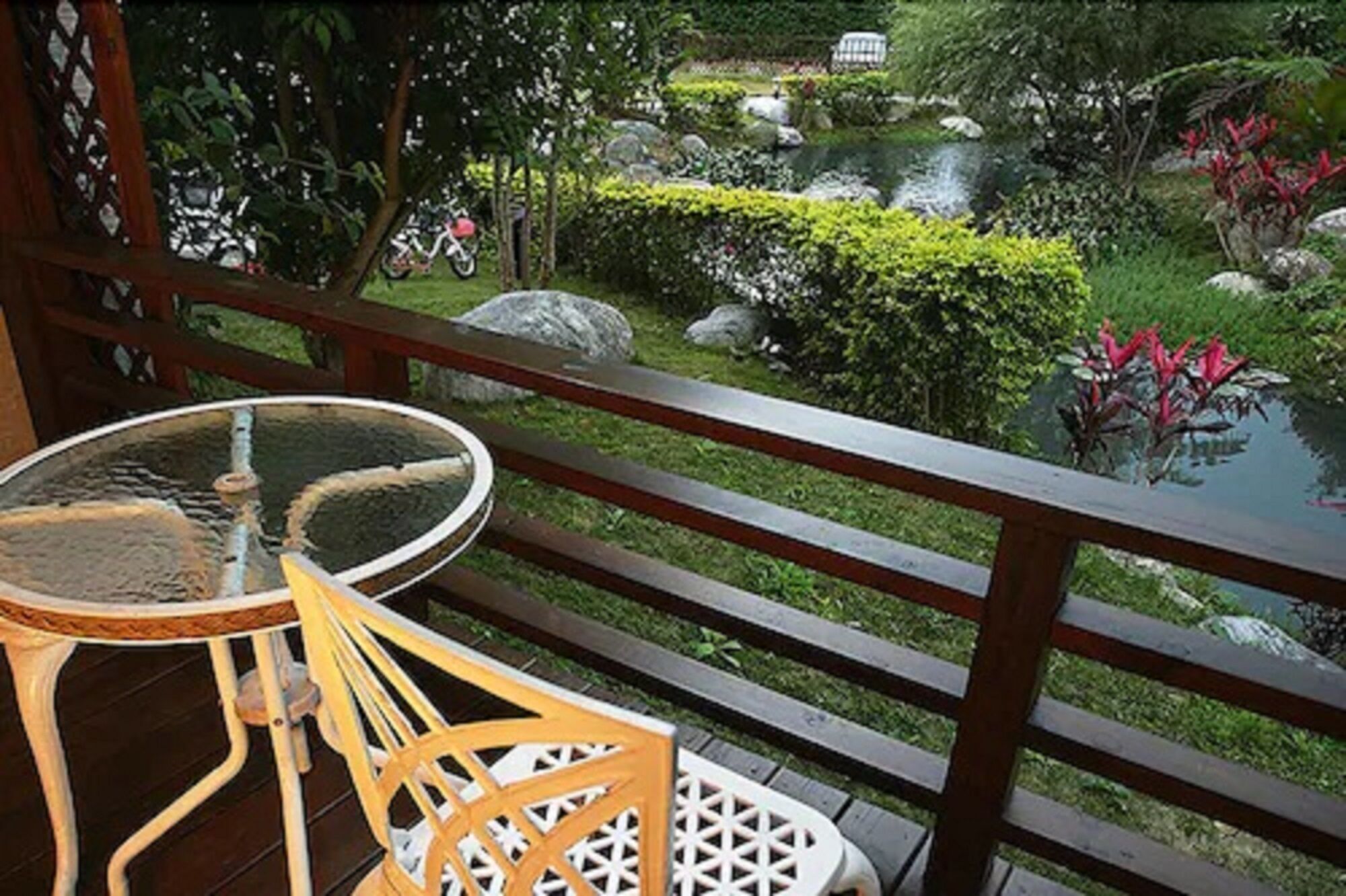 Terraza de madera con una mesa de vidrio y dos sillas, con vistas a un jardín que tiene un estanque y diversas plantas.