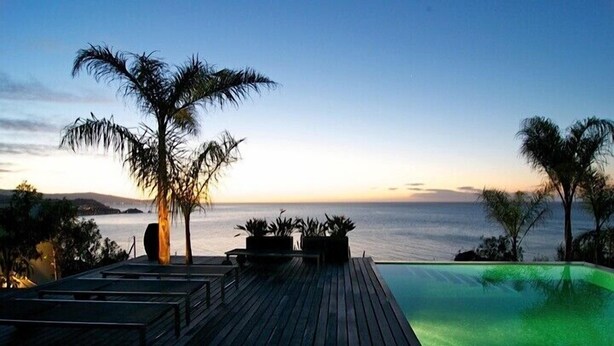 Une terrasse en bois avec une piscine, des palmiers et une vue sur la mer au coucher du soleil.