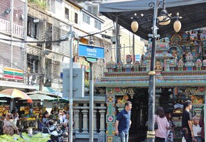 Calle concurrida con la entrada colorida de un templo, un cartel que indica 'Thanon Pan' y una zona de mercado con gente y puestos.