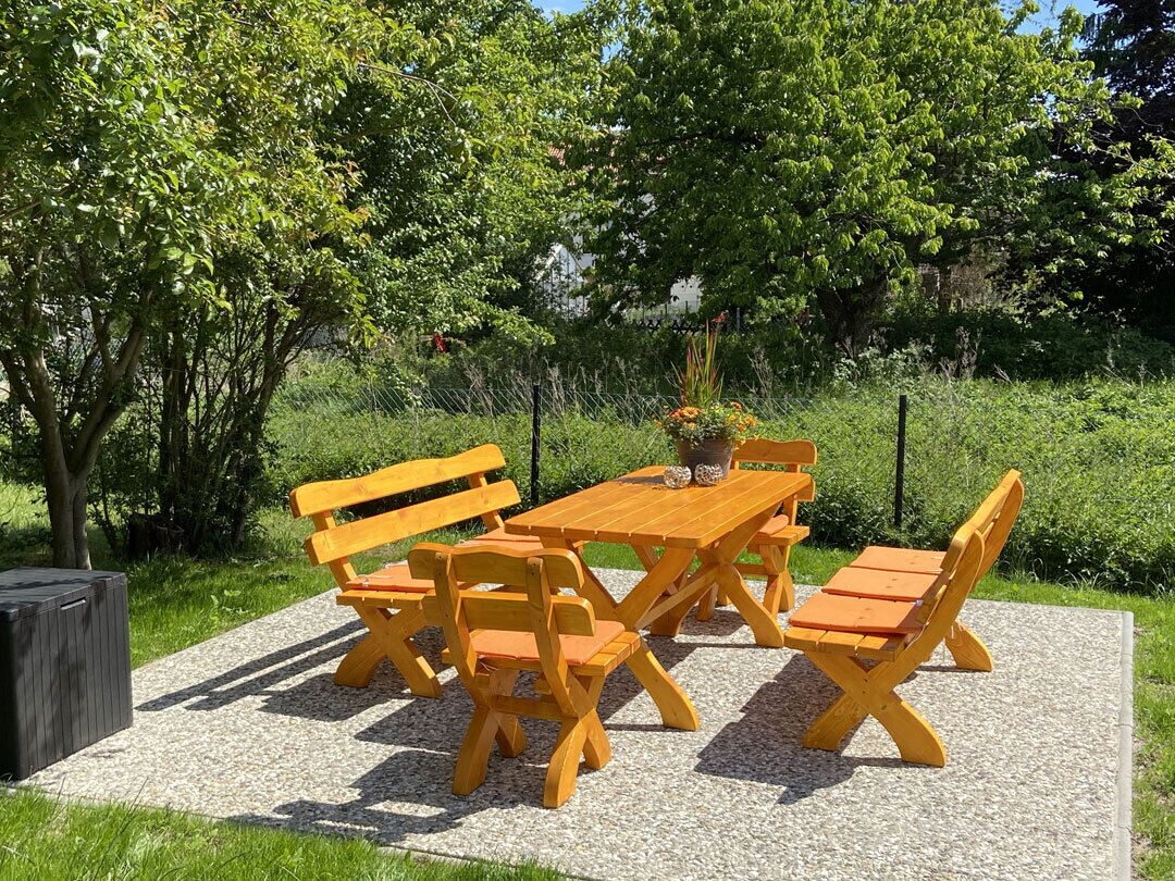 Un espace extérieur aménagé avec des sièges en bois, comprenant une table et des bancs, le tout entouré de verdure et d’arbres.