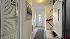 Un couloir étroit, avec une porte au fond, un radiateur, une petite table recouverte d’une nappe à motifs et une chaise rouge.