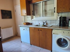 Una cocina compacta con lavavajillas, fregadero, armarios de madera, ventana con persianas y lavadora.