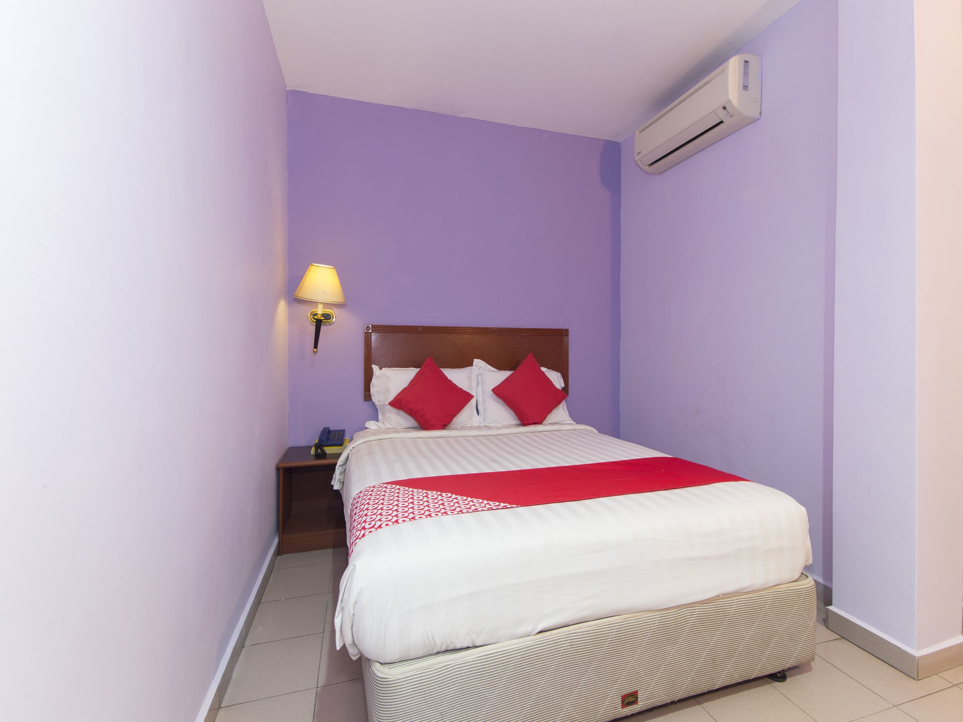 A small, neatly arranged bedroom with a single bed, a bedside lamp, a nightstand, and an air conditioning unit on the wall.