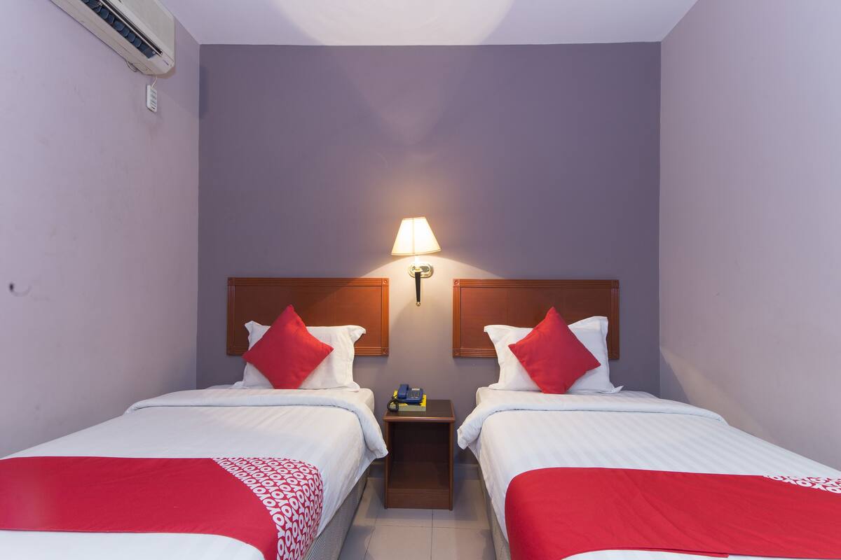 A hotel room with two beds, each with a red and white bedspread, a nightstand with a lamp, and an air conditioning unit on the wall.